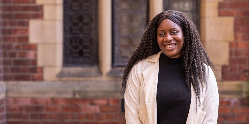 A student standing on campus in front of a red brick building and smiling.